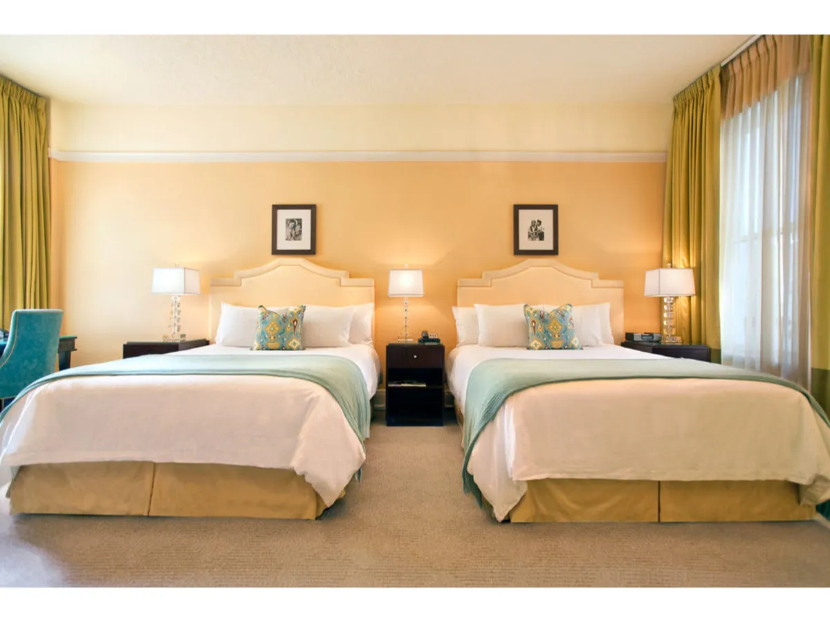 The image showcases a serene and stylish hotel room featuring two plush queen beds adorned with decorative pillows, elegant nightstands with softly glowing lamps, and tasteful artwork on the walls. The ambiance is calm and inviting, perfect for relaxing, reading, or enjoying time with loved ones.