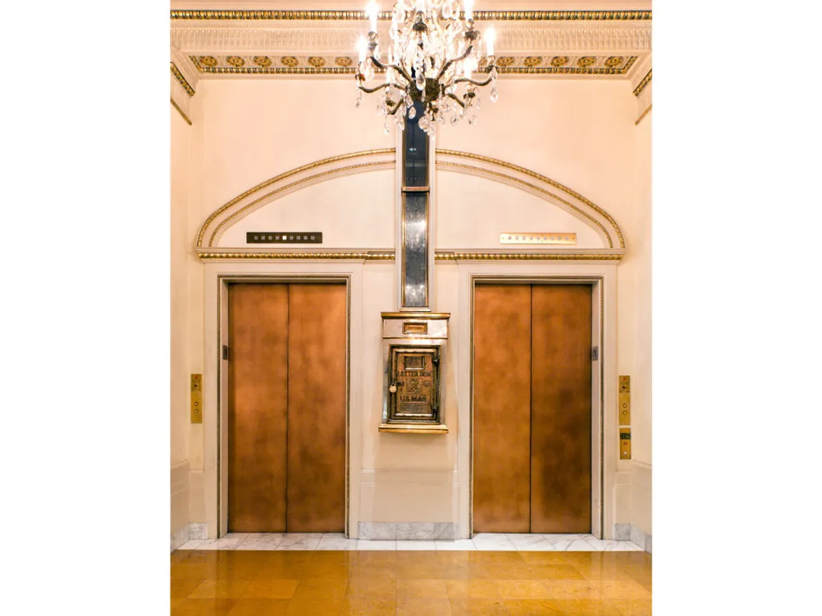 A luxurious hotel lobby scene featuring ornate wall details, a sparkling chandelier, and elevator doors. The setting feels elegant and welcoming, with guests waiting for the elevator and engaging in casual conversation before heading out.