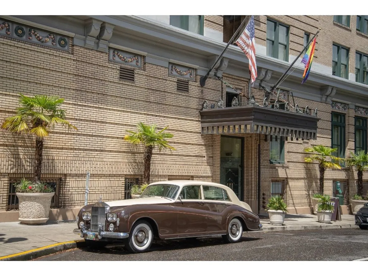 An inviting exterior shot featuring a classic car at the building’s entrance, framed by palm trees and a retro awning. The scene exudes timeless elegance and the anticipation of arriving guests, with architectural details hinting at the property’s boutique character.