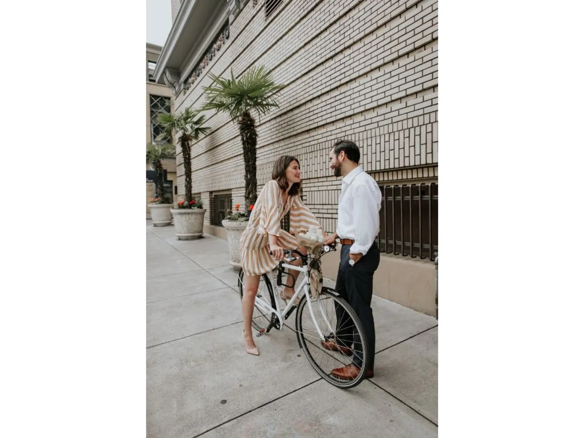 An exterior-adjacent scene showing a couple interacting by bicycles, with potted plants and a textured brick wall in the background. The atmosphere is playful and romantic, suggesting urban exploration and the availability of complimentary bike rentals for guests.