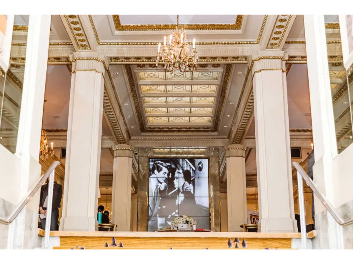 This luxurious interior scene showcases an ornate chandelier, an intricately designed ceiling, and a large screen displaying a black-and-white image. The space is accented with a decorative floral arrangement, creating a lavish setting ideal for formal gatherings and art exhibitions.