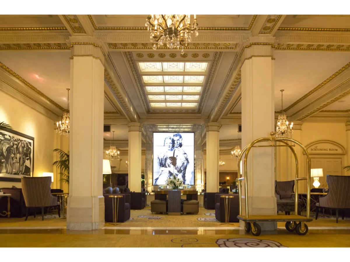 An elegant room anchored by a grand chandelier, a dramatic mural, and a thoughtfully arranged seating area. A luggage cart in the foreground hints at the space's adaptability for both leisure and business uses, making it suitable for meetings, networking, or relaxation.
