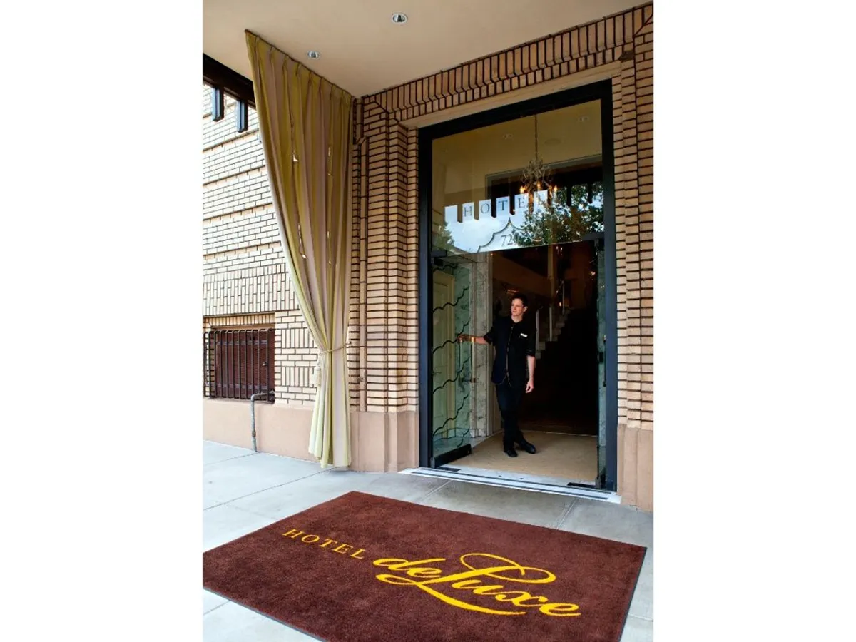 A friendly staff member greets guests at the hotel's entrance, with the hotel mat and entrance door visible. The scene conveys a sense of personalized hospitality and smooth arrivals, highlighting the importance of comfort and assistance for all visitors.