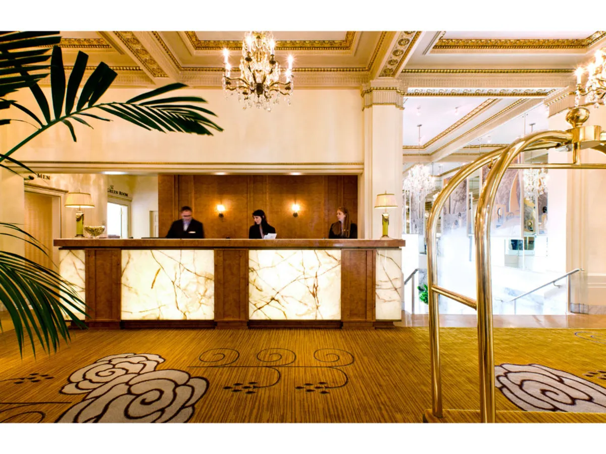 A view of the hotel's reception desk bathed in warm light, with staff members visible behind the counter ready to assist. Chandeliers hang from the ceiling, and a decorative plant adds a welcoming touch to the space. The scene exudes luxury and provides a glimpse into the attentive service and inviting atmosphere upon arrival.