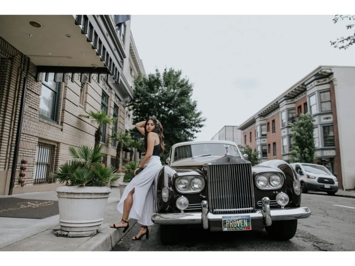 This exterior image captures a vintage car parked near an urban building facade, with a woman in stylish attire standing nearby. The background features classic architectural elements, suggesting a lively, sophisticated city environment ideal for social gatherings or casual strolls.