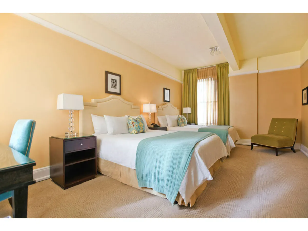 A bright, inviting bedroom with two queen beds dressed in light-colored bedding, accented with turquoise throws and patterned pillows. The room features modern bedside lamps, a small desk and chair, and a plush armchair, all arranged with generous space between them for easy navigation.