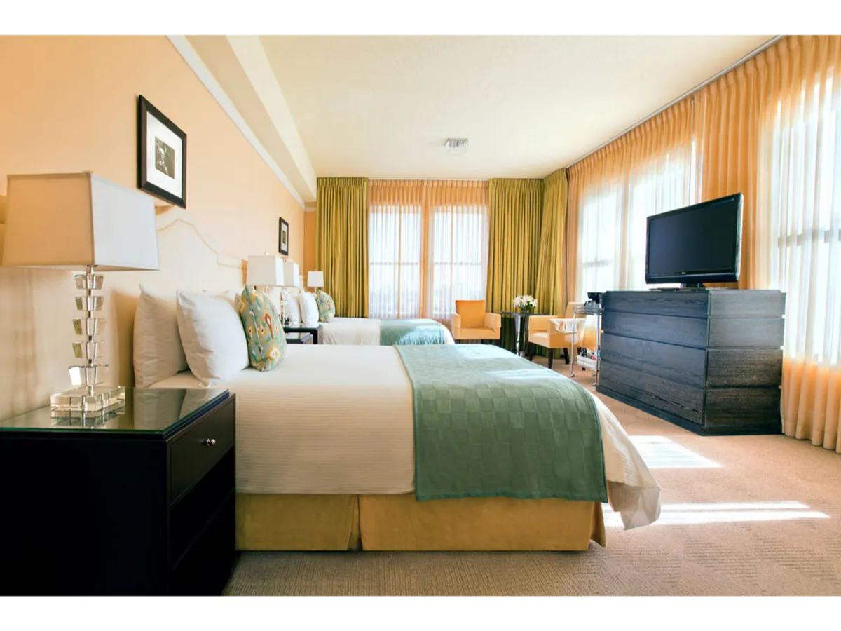 The image displays a tranquil, welcoming bedroom featuring two beds adorned with colorful bedding, large windows draped with sheer curtains allowing natural light, a television unit, and a cozy seating area. The layout is open and uncluttered, providing ample clear floor space ideal for ease of movement and accessibility.