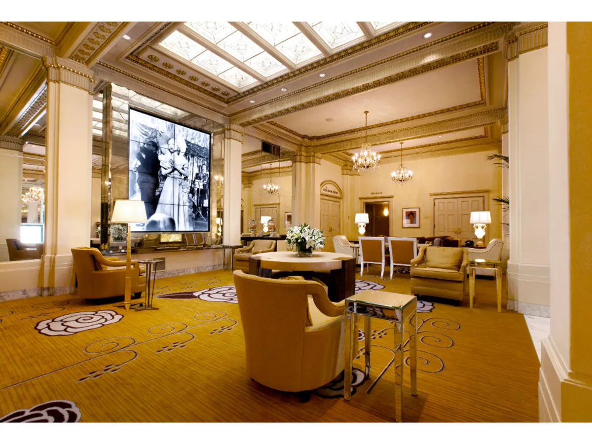 A refined common area featuring a large digital screen displaying a historical black and white photograph, elegant chandeliers, stylish seating, and a prominent floral centerpiece on a table. The ambiance is luxurious and inviting, highlighting both the curated art collection and historic charm.