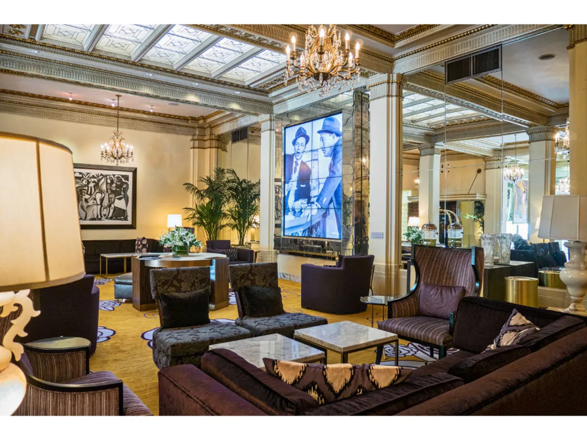 A plush, sophisticated interior with a chandelier, large screen displaying historical imagery, and an inviting seating arrangement surrounded by decorative plants. The space is designed for social interactions and art appreciation, with an ambiance suited for events or casual gatherings.